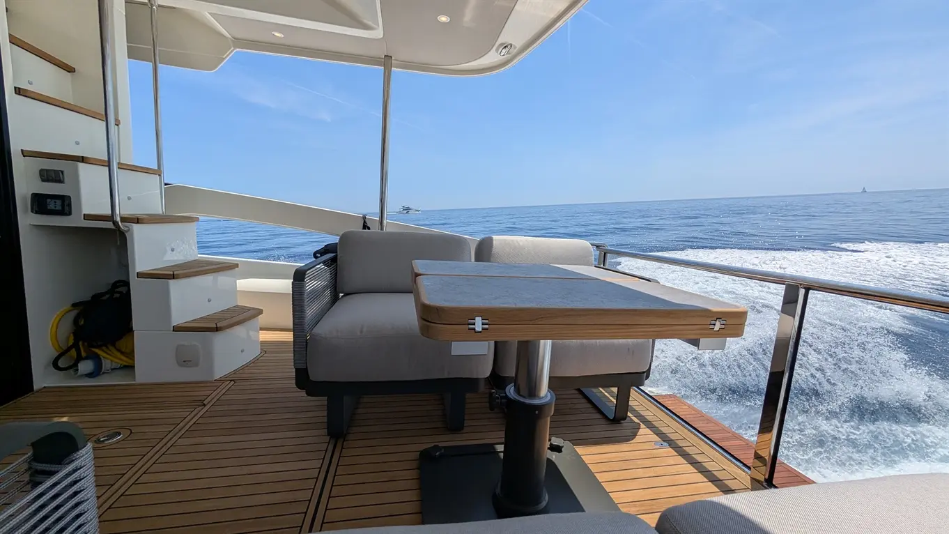 A stylish outdoor seating area on a yacht, featuring a table and two armchairs, with ocean views and blue skies.
